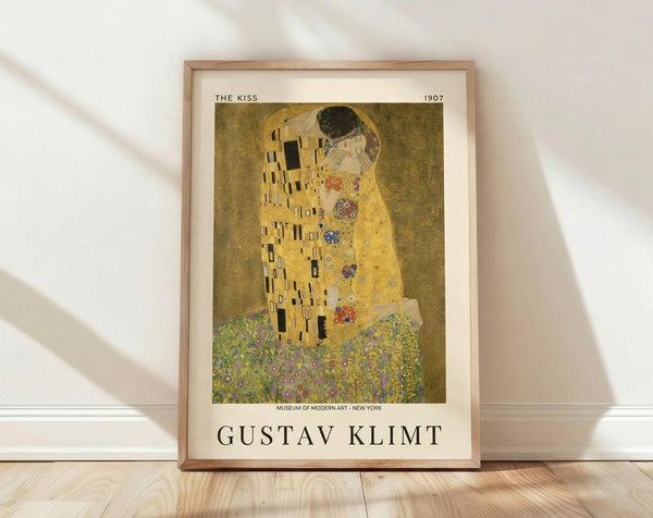 May include: Framed poster print of Klimt's famous golden painting showing an embracing couple against ornate patterned robes, displayed on light wooden floor against white textured wall with natural window lighting creating soft shadows across the scene.
