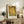 May include: Framed reproduction of Klimt's famous golden painting showing two embracing figures in ornate robes, displayed on rustic wooden sideboard beside fern plant, ceramic vase, and stack of books in neutral-toned living room with beige chair and pillows