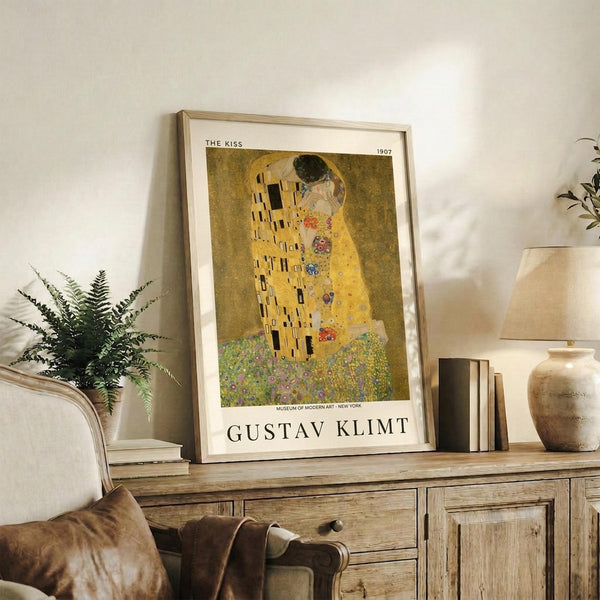 May include: Framed reproduction of Klimt's famous golden painting showing two embracing figures in ornate robes, displayed on rustic wooden sideboard beside fern plant, ceramic vase, and stack of books in neutral-toned living room with beige chair and pillows