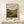 May include: Framed landscape poster print in thin wood frame displayed on light wooden shelf against white wall. Artwork shows rolling hills in earth tones with winding river, distant factory buildings, stylized trees, and cloudy sky in vintage print style. Small white wrapped gift box with natural twine sits on shelf beside the frame.