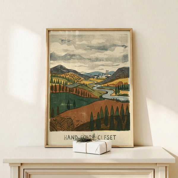 May include: Framed landscape poster print in thin wood frame displayed on light wooden shelf against white wall. Artwork shows rolling hills in earth tones with winding river, distant factory buildings, stylized trees, and cloudy sky in vintage print style. Small white wrapped gift box with natural twine sits on shelf beside the frame.