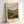May include: A framed print leaning against a white wall showing a stylized landscape with rolling hills in green and orange tones, a winding river, dark mountains, and cloudy sky. The artwork features a woodblock or linocut aesthetic with bold, simplified forms and earthy colors. Text at bottom reads "HAND FLORA OFFSET" in block letters. The print is displayed in a light wooden frame on what appears to be a white surface.