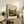 May include: A framed landscape print featuring rolling hills in earth tones of green, brown, and orange with a winding river, stylized trees, and cloudy sky. The artwork sits on a rustic wooden sideboard beside a fern plant, ceramic vase, stacked books, and brown leather chair in a cozy living room setting with neutral walls.