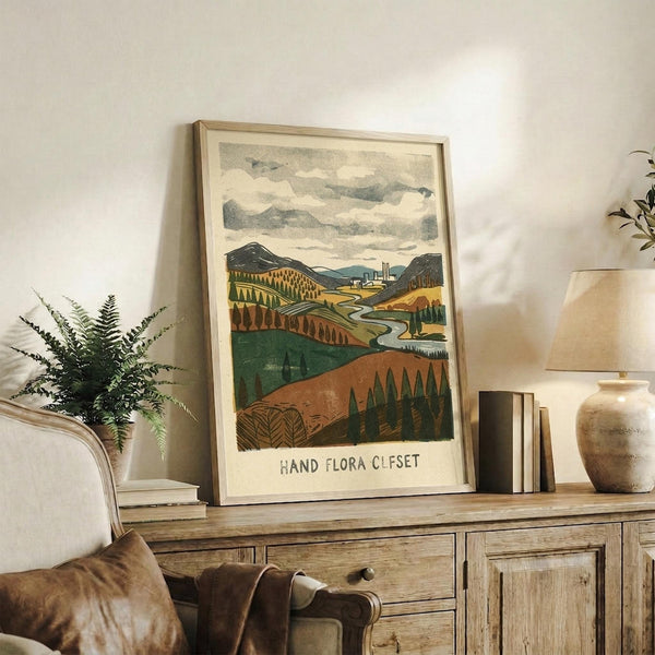 May include: A framed landscape print featuring rolling hills in earth tones of green, brown, and orange with a winding river, stylized trees, and cloudy sky. The artwork sits on a rustic wooden sideboard beside a fern plant, ceramic vase, stacked books, and brown leather chair in a cozy living room setting with neutral walls.