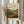 May include: Person in beige ruffled sleeves holding framed landscape print showing rolling hills, winding river, distant industrial buildings, and cloudy sky in muted greens, browns, and yellows. Light wooden frame against white curtained window with dried pampas grass stems visible to the left.