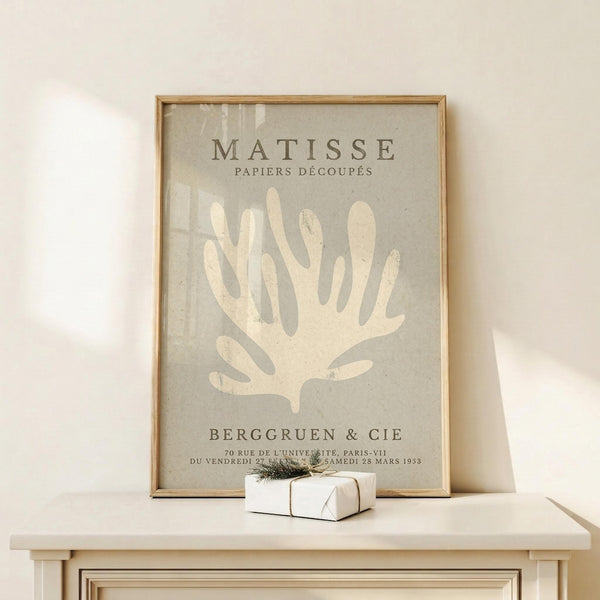 May include: Framed vintage-style art print featuring cream-colored coral or seaweed design on sage green background with "MATISSE PAPIERS DÉCOUPÉS" and "BERGGRUEN & CIE" text, displayed in thin gold frame on light wood console table next to small wrapped white gift with twine and greenery sprig against cream wall