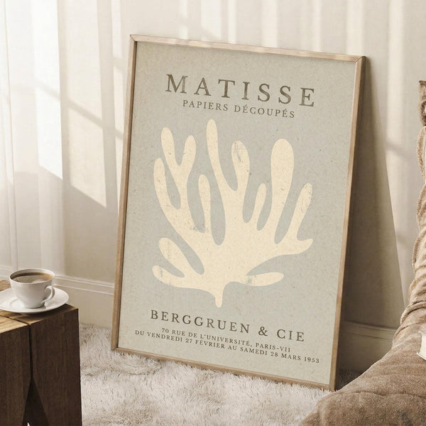 May include: Framed beige and cream Matisse exhibition poster featuring abstract coral-like botanical form, displayed leaning against white wall corner on textured white rug. Light wood frame, soft natural lighting creates geometric shadows. Coffee cup on wooden block visible to left, cozy neutral interior setting.