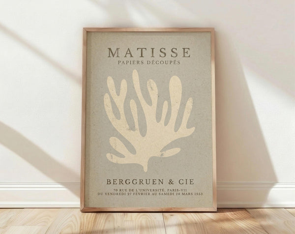 May include: Framed poster with beige background featuring cream-colored coral or seaweed silhouette in Matisse's cut-out style. Text reads "MATISSE PAPIERS DÉCOUPÉS" at top and "BERGGRUEN & CIE" with Paris gallery details below. Natural wood frame sits on light wooden floor against white wall with soft shadows.