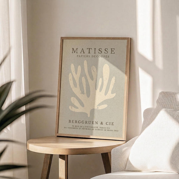 May include: Framed art print featuring cream-colored coral silhouette on sage green background with "MATISSE PAPIERS DÉCOUPÉS" and "BERGGRUEN & CIE" text, displayed on round wooden side table next to beige upholstered chair with plant leaves visible, sunlight casting shadows across neutral-toned room setting