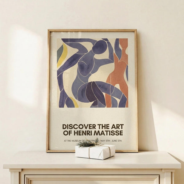 May include: Framed poster featuring abstract dancing figures in purple, blue, orange and yellow cut-out style shapes against cream background, displayed on light wood console table beside small wrapped gift with twine and greenery sprig against white wall