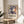 May include: Framed poster with abstract blue cut-out style figures dancing, featuring flowing organic shapes in navy blue, coral orange, and yellow on cream background. Exhibition-style text below reads "Discover the Art of Henri Matisse" with museum details. Wooden frame sits on round wooden side table next to beige upholstered chair, with plant leaves visible and natural sunlight casting shadows on white wall.