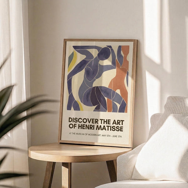 May include: Framed poster with abstract blue cut-out style figures dancing, featuring flowing organic shapes in navy blue, coral orange, and yellow on cream background. Exhibition-style text below reads "Discover the Art of Henri Matisse" with museum details. Wooden frame sits on round wooden side table next to beige upholstered chair, with plant leaves visible and natural sunlight casting shadows on white wall.