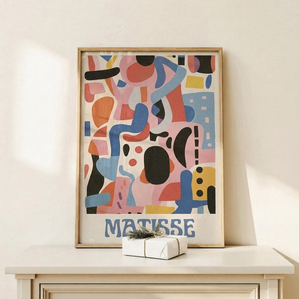May include: Framed abstract art print in light wood frame featuring colorful geometric shapes in pink, blue, yellow, orange and black with "MATISSE" text at bottom, displayed on white wooden shelf next to small wrapped gift with twine and greenery sprig against cream wall