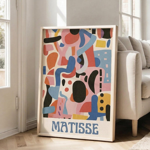 May include: Colorful abstract art poster with organic shapes in pink, blue, orange, yellow, and black, featuring "MATISSE" text at bottom, displayed in light wood frame leaning against white wall next to beige couch in bright living room with large windows and natural light