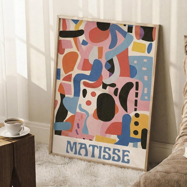 May include: Framed abstract art poster with colorful geometric shapes in pink, blue, orange, yellow and black, featuring "MATISSE" text at bottom. Light wood frame leaning against white wall corner on fluffy white rug, with coffee cup on wooden block and beige blanket visible in cozy room setting.