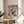 May include: Colorful abstract art poster with geometric shapes in pink, blue, orange, and black with "MATISSE" text at bottom, displayed in thin wooden frame on round wooden side table next to beige upholstered chair. Sunlight casts shadows across white wall behind artwork. Green plant leaves visible in corner of minimalist modern room setting.