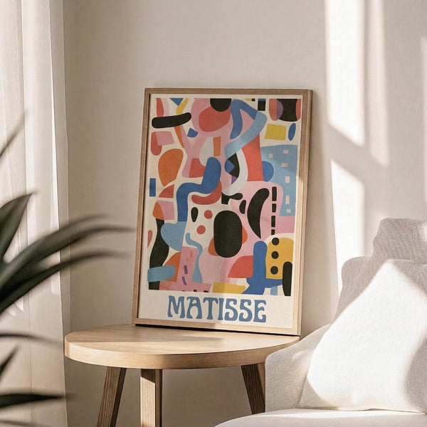 May include: Colorful abstract art poster with geometric shapes in pink, blue, orange, and black with "MATISSE" text at bottom, displayed in thin wooden frame on round wooden side table next to beige upholstered chair. Sunlight casts shadows across white wall behind artwork. Green plant leaves visible in corner of minimalist modern room setting.