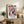 May include: Colorful abstract art poster with geometric shapes in pink, blue, orange, and black, displayed in light wood frame on rustic wooden sideboard. Room setting shows fern plant, books, ceramic vase, and beige armchair with brown leather cushion against cream wall.