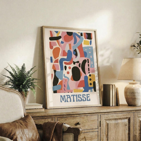 May include: Colorful abstract art poster with geometric shapes in pink, blue, orange, and black, displayed in light wood frame on rustic wooden sideboard. Room setting shows fern plant, books, ceramic vase, and beige armchair with brown leather cushion against cream wall.