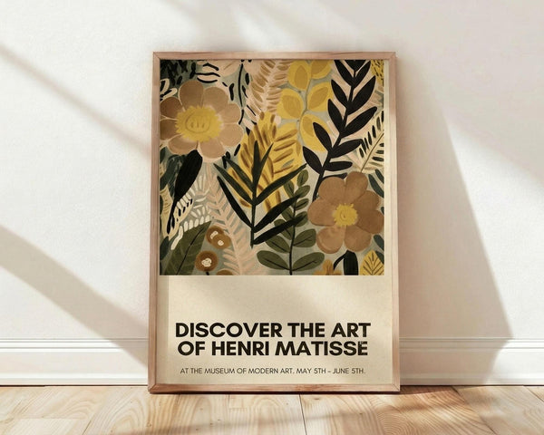 May include: Framed poster with colorful botanical illustration featuring stylized flowers and leaves in yellow, brown, green, and black tones. Exhibition-style text below reads "DISCOVER THE ART OF HENRI MATISSE" with museum and date details. Light wood frame rests against white wall on natural wood flooring with soft shadows cast across the scene.