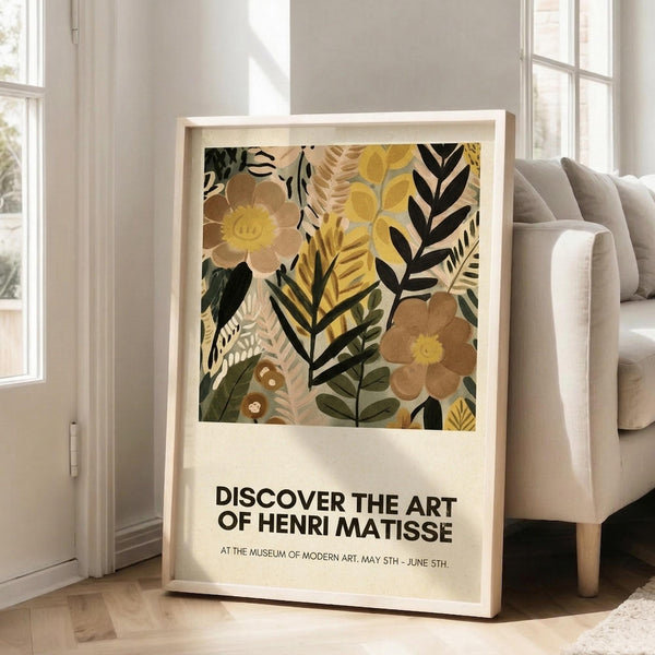May include: Framed botanical art print with colorful abstract flowers and leaves in yellow, brown, green, and black tones, displayed on light wooden floors against white walls next to a beige sofa. The poster features exhibition text about Henri Matisse and shows layered floral collage elements with various leaf patterns and blooms in an earthy, vintage-inspired color palette.