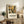 May include: Framed botanical art poster with colorful flowers and leaves in yellow, brown, and green tones, displayed on rustic wooden sideboard alongside fern plant, ceramic vase, and books in neutral living room setting with beige chair and warm lighting.