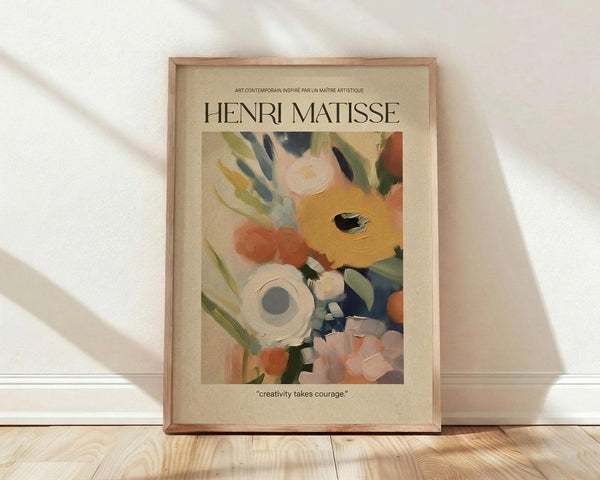 May include: Framed Henri Matisse poster print leaning against white wall on light wood floor. Abstract floral artwork features bold brushstrokes in orange, pink, blue, green and white with flowing organic shapes. Cream matting surrounds colorful botanical design. Quote "creativity takes courage" appears below image. Natural light casts diagonal shadows across scene.