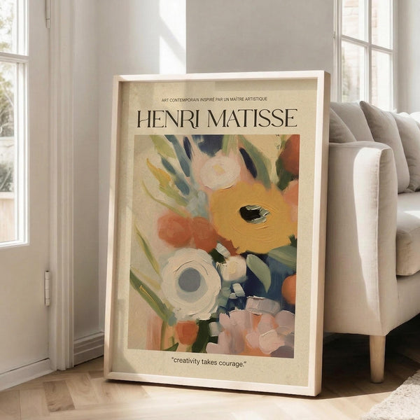 May include: Framed art print featuring colorful floral painting with orange, pink, white and blue blooms against cream background. Henri Matisse text at top with "creativity takes courage" quote below. Light wood frame leaning against white wall next to beige upholstered chair in bright, minimalist living room with hardwood floors and large windows.