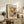 May include: Framed Henri Matisse art print displayed on rustic wooden sideboard in cozy living room setting. The colorful abstract floral artwork features vibrant yellows, oranges, pinks and blues with white flowers. Scene includes fern plant, stacked books, ceramic vase, brown leather chair, and warm natural lighting creating shadows on cream walls.