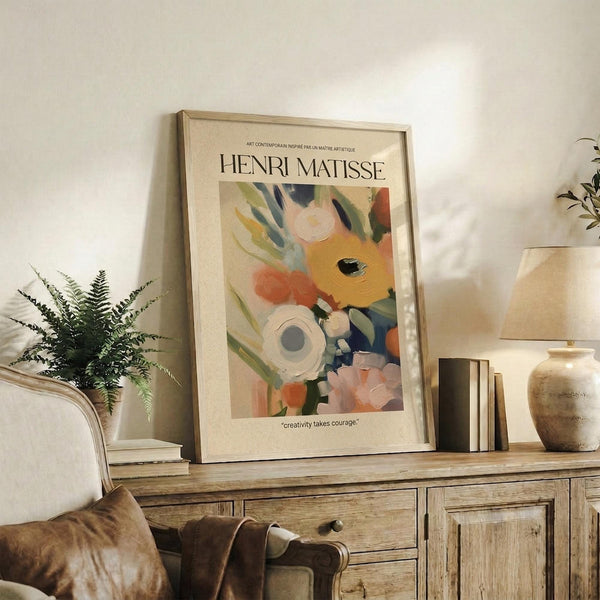 May include: Framed Henri Matisse art print displayed on rustic wooden sideboard in cozy living room setting. The colorful abstract floral artwork features vibrant yellows, oranges, pinks and blues with white flowers. Scene includes fern plant, stacked books, ceramic vase, brown leather chair, and warm natural lighting creating shadows on cream walls.