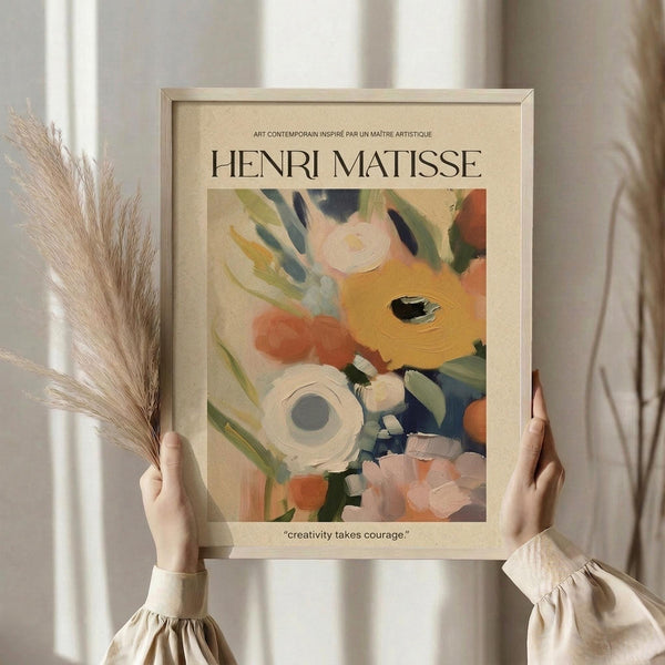 May include: Person in cream sweater holding framed Henri Matisse-inspired floral art print featuring abstract flowers in soft oranges, pinks, greens, and blues with "creativity takes courage" quote below. Dried pampas grass visible on left side against neutral background with natural window lighting.