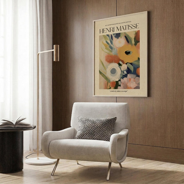 May include: Cream-colored Henri Matisse poster with abstract floral design in orange, yellow, green and blue tones, displayed in light wood frame on textured wood wall panel. Modern beige armchair with geometric patterned pillow sits below artwork. Gold floor lamp and dark side table with open book complete the warm, contemporary interior scene.