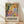 May include: Framed poster with colorful abstract artwork featuring vibrant fruits, flowers, and foliage in yellows, oranges, greens, blues and pinks with black tree branches. Exhibition-style poster with bold black text reading "DISCOVER THE ART OF HENRI MATISSE" and museum details below. Natural wood frame leaning against white wall on light wooden floor with soft shadows.