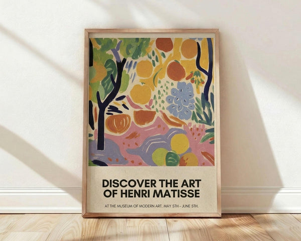 May include: Framed poster with colorful abstract artwork featuring vibrant fruits, flowers, and foliage in yellows, oranges, greens, blues and pinks with black tree branches. Exhibition-style poster with bold black text reading "DISCOVER THE ART OF HENRI MATISSE" and museum details below. Natural wood frame leaning against white wall on light wooden floor with soft shadows.
