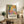 May include: Colorful Henri Matisse exhibition poster in light wood frame, leaning against white wall on rustic wooden console. Artwork features vibrant fruit, flowers and foliage in yellow, orange, green and purple with black tree branches. Room styled with fern plant, cream lamp, ceramic vase, stacked books and brown leather chair cushions.