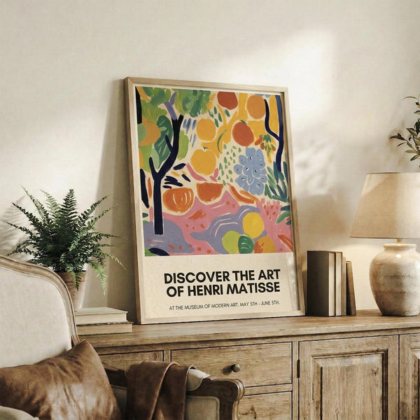 May include: Colorful Henri Matisse exhibition poster in light wood frame, leaning against white wall on rustic wooden console. Artwork features vibrant fruit, flowers and foliage in yellow, orange, green and purple with black tree branches. Room styled with fern plant, cream lamp, ceramic vase, stacked books and brown leather chair cushions.