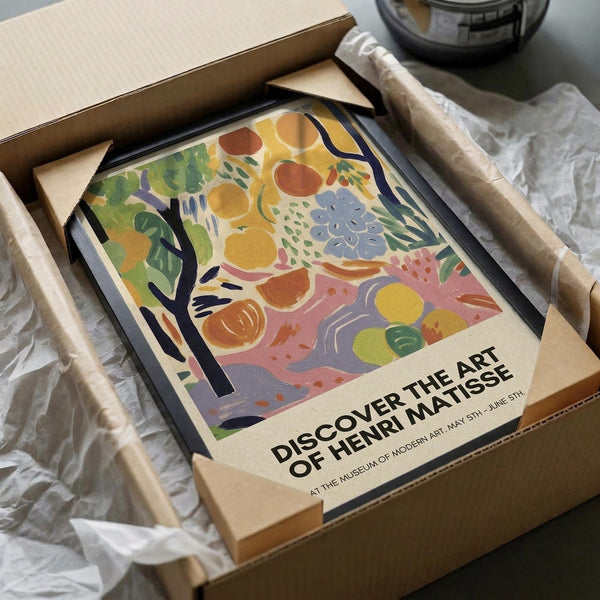 May include: Colorful Henri Matisse-style art print featuring abstract fruits, flowers and foliage in vibrant yellows, greens, blues, pinks and oranges, displayed in black frame inside cardboard shipping box with protective tissue paper wrapping on white surface