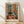 May include: Framed Henri Matisse art print leaning against white wall on light wood floor. The colorful fauve-style interior painting shows an orange and blue bedroom with open French doors, wooden bed, small round stool, and black and white cat on terracotta floor tiles. Natural light creates soft shadows on the wall behind the light wood frame.