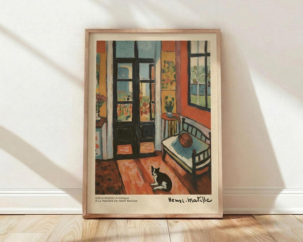 May include: Framed Henri Matisse art print leaning against white wall on light wood floor. The colorful fauve-style interior painting shows an orange and blue bedroom with open French doors, wooden bed, small round stool, and black and white cat on terracotta floor tiles. Natural light creates soft shadows on the wall behind the light wood frame.