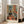May include: Framed art print leaning against white wall beside beige sofa in bright room with windows. Artwork shows colorful interior scene with black and white cat on wooden floor, green doors, orange walls, small bed, and potted plants. Matisse-inspired style with vibrant blues, oranges, and greens in light wood frame.