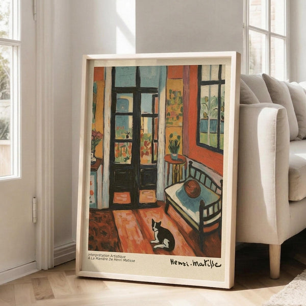 May include: Framed art print leaning against white wall beside beige sofa in bright room with windows. Artwork shows colorful interior scene with black and white cat on wooden floor, green doors, orange walls, small bed, and potted plants. Matisse-inspired style with vibrant blues, oranges, and greens in light wood frame.