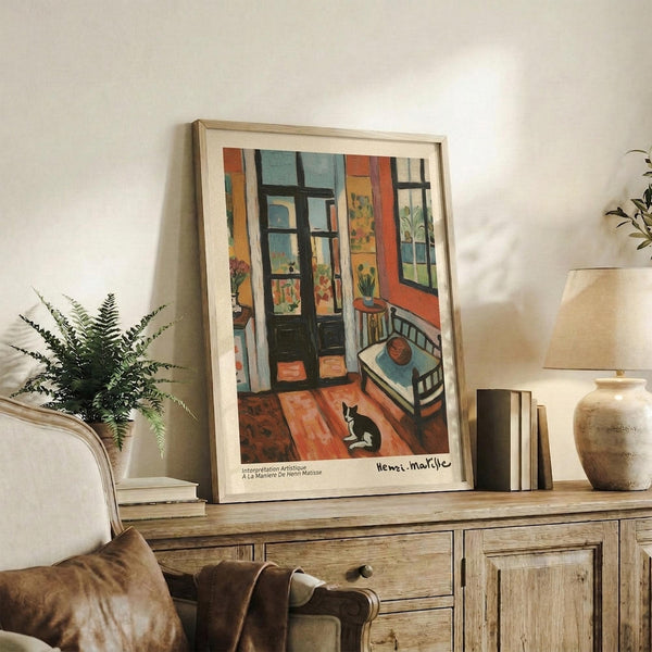 May include: Framed Henri Matisse reproduction poster showing colorful interior scene with cat on wooden floor, displayed on rustic wooden sideboard alongside ceramic vase, books, and fern plant in cozy living room setting with cream upholstered chair and neutral walls