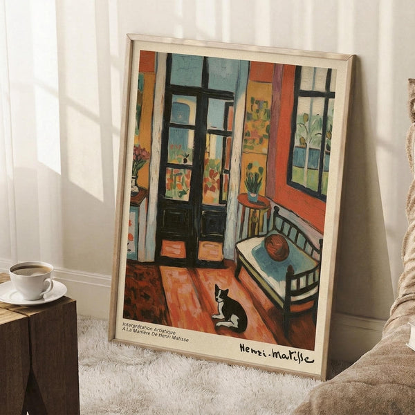 May include: Framed Henri Matisse art print showing colorful interior scene with black and white cat on orange wooden floor, green chair, bed, and French doors opening to bright rooms beyond. Print features vibrant blues, oranges, and greens in Matisse's distinctive style. Frame leans against white wall next to coffee cup on wooden block and textured throw blanket in cozy room setting.