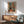 May include: Framed Henri Matisse interior scene print hung above wooden sideboard with white lamp. Artwork depicts colorful room with black and white cat, furniture, windows, and warm orange, blue, and green tones. Mid-century modern living room setting with sage green upholstered chair, round coffee table, and natural sunlight casting shadows on white wall.
