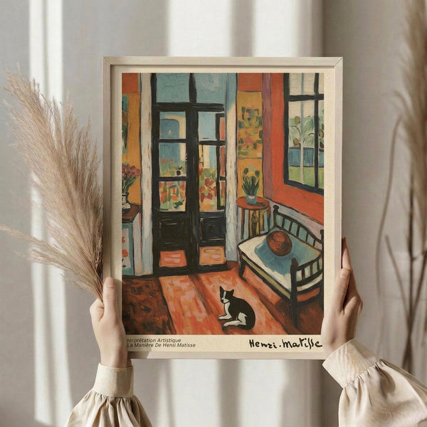 May include: Person holding framed Henri Matisse-style interior scene artwork showing colorful room with black and white cat on wooden floor, single bed, open French doors, potted plants, warm oranges and blues. Light wooden frame, dried wheat stems visible, soft natural lighting against white curtained background.