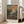 May include: Framed art print in light wood frame leaning against white wall next to beige sofa. Artwork shows impressionist landscape with dark green foliage at bottom, colorful sky with blue, pink, coral, and yellow brushstrokes above. Small figures visible among the vegetation. Text reads "Henri Matisse Paris" at bottom. Bright natural light from windows creates soft shadows on hardwood floor.