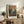 May include: Framed Henri Matisse art print displayed on rustic wooden credenza in warm-toned living room. Poster features vibrant landscape with colorful sky in blues, oranges and pinks above lush green foliage and small figures below. Natural wood frame with white matting. Scene includes fern plant, leather chair, ceramic vase, stacked books and soft natural lighting.