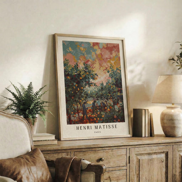 May include: Framed Henri Matisse art print displayed on rustic wooden credenza in warm-toned living room. Poster features vibrant landscape with colorful sky in blues, oranges and pinks above lush green foliage and small figures below. Natural wood frame with white matting. Scene includes fern plant, leather chair, ceramic vase, stacked books and soft natural lighting.
