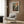 May include: Henri Matisse poster print in light wood frame hanging on wood-paneled wall above cream upholstered armchair with geometric patterned pillow. The colorful artwork depicts an orchard scene with figures among fruit trees under a vibrant sky in impressionistic style. Modern brass floor lamp and dark side table with open book complete the contemporary reading nook setting.