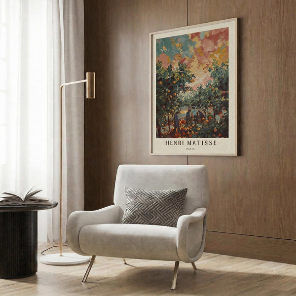 May include: Henri Matisse poster print in light wood frame hanging on wood-paneled wall above cream upholstered armchair with geometric patterned pillow. The colorful artwork depicts an orchard scene with figures among fruit trees under a vibrant sky in impressionistic style. Modern brass floor lamp and dark side table with open book complete the contemporary reading nook setting.